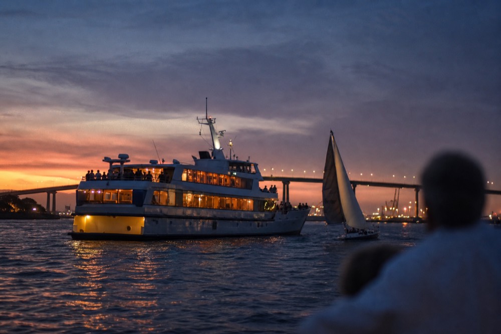 Lit dinner cruise ship and a small sunset sail crossing paths at dusk on San Diego Bay with bridge lights behind