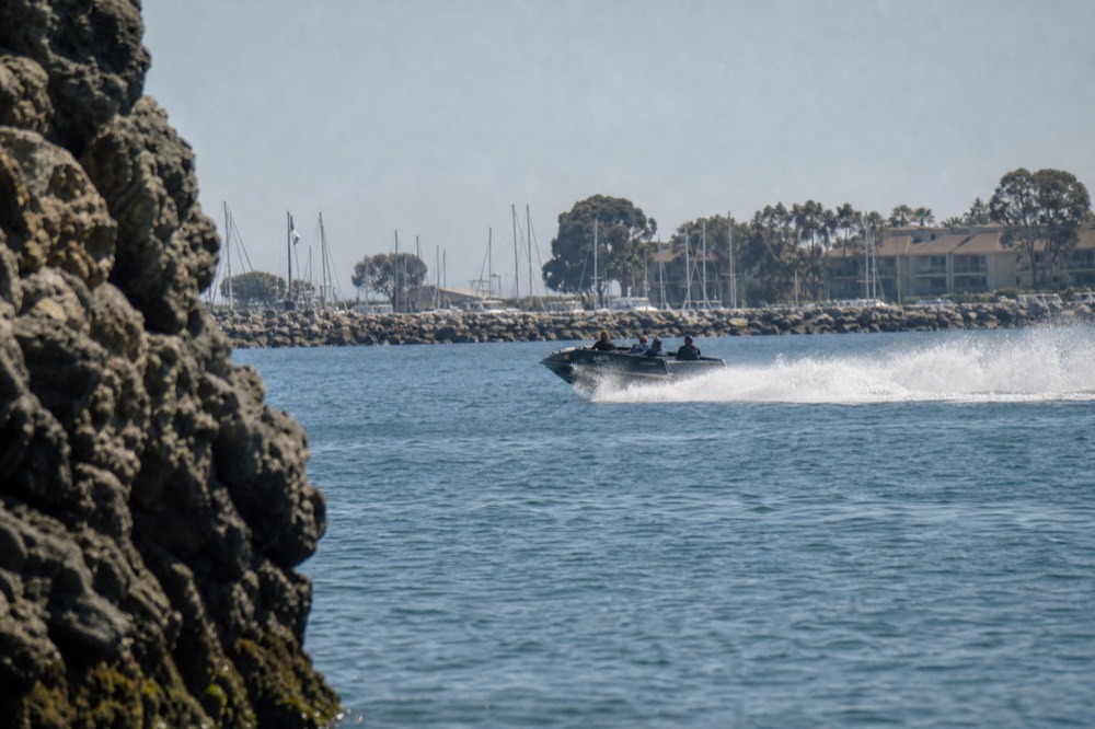 A jet boat at full speed with a spray fan on flat harbor water, late-morning light
