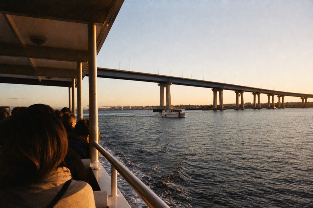 The small Coronado passenger ferry crossing San Diego Bay toward the bridge at golden hour, long shadows and warm low light