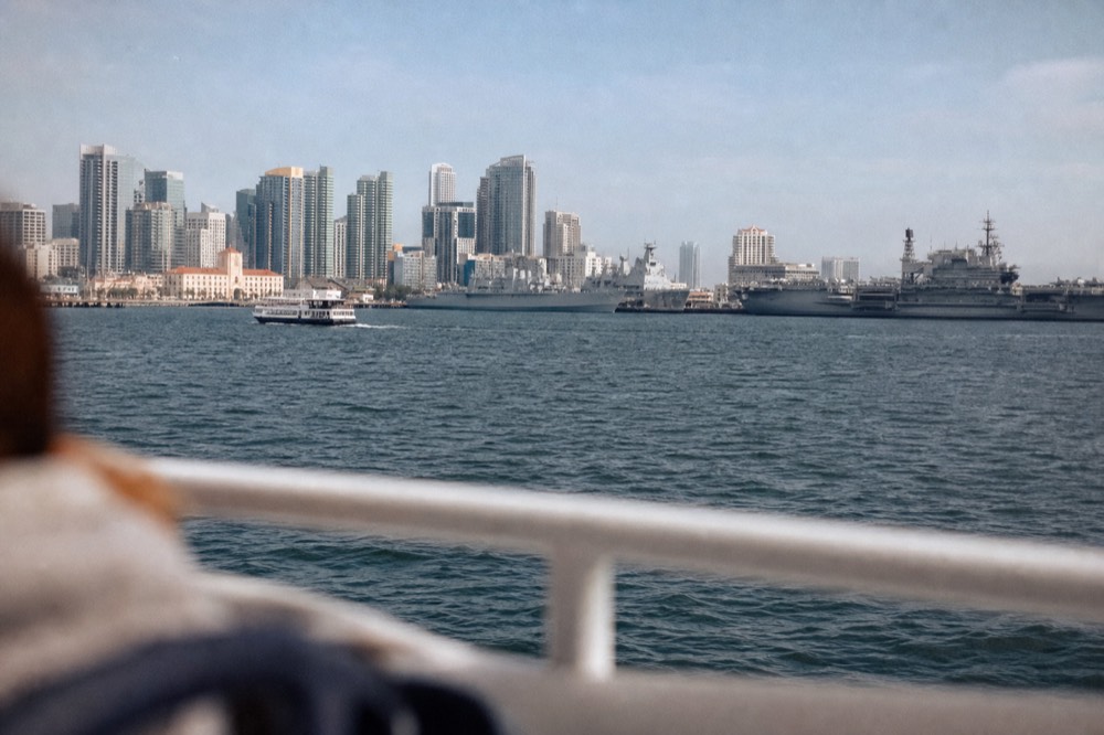 The San Diego harbor cruise route with bay water, gray naval ships and the city skyline, a railing or shoulder softly out of focus in the fo