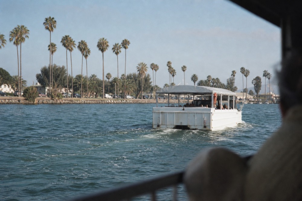 A boxy amphibious sightseeing vehicle around San Diego's palm-lined waterfront, a railing or shoulder softly out of focus in the foreground