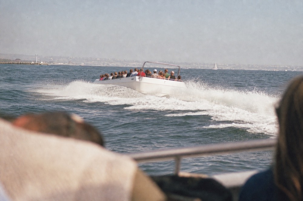 A fast open jet boat throwing spray on San Diego Bay, a railing or shoulder softly out of focus in the foreground