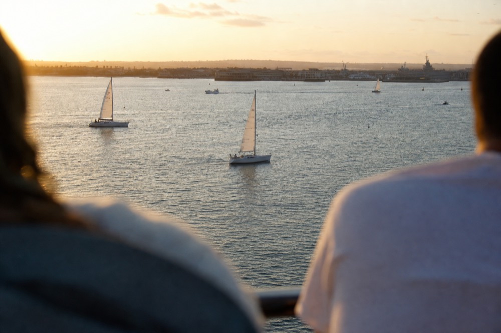 Sunset light over San Diego Bay and its sailboats, a railing or shoulder softly out of focus in the foreground